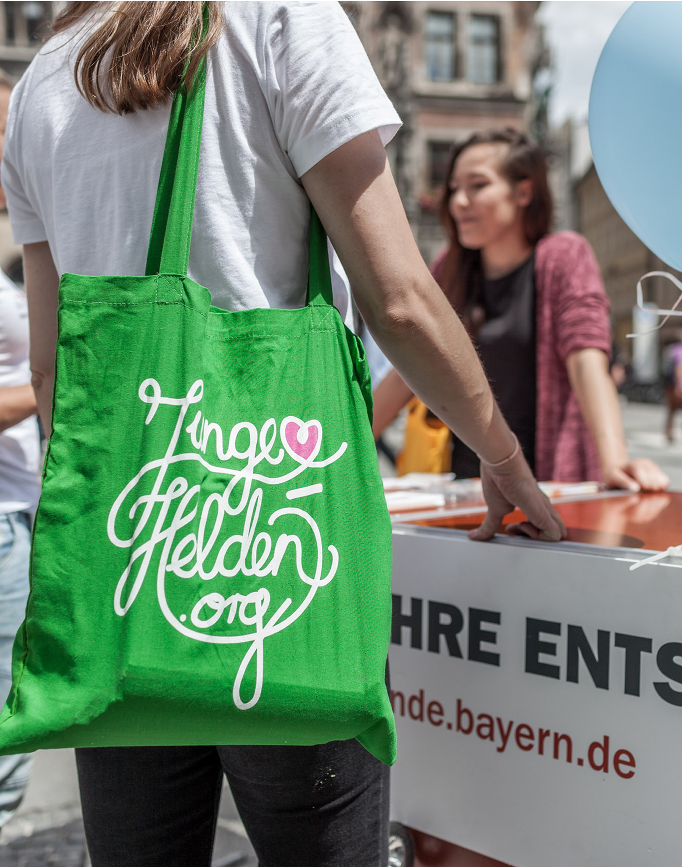 Woman with green "Young Heroes" bag at the information desk of the Young Heroes (Photo)